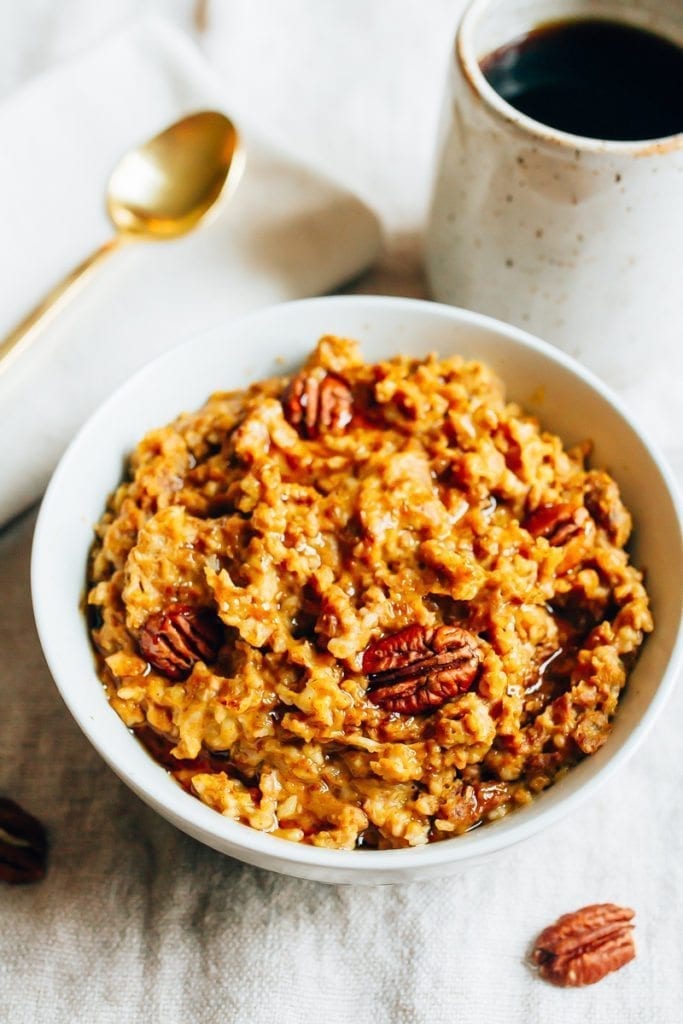 Slow Cooker Pumpkin Pie Oatmeal in a bowl, topped with pecans. A spoon, napkin and mug of coffee to the side.