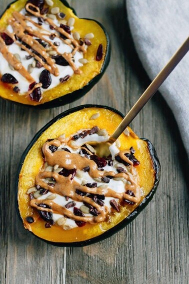 2 acorn squash breakfast bowls with a spoon, filled with yogurt, sunflower seeds, dried cranberries, and drizzled with peanut butter.