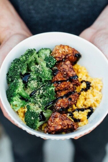 Rice bowl with a row of tempeh and broccoli in a white bowl in someones hands.