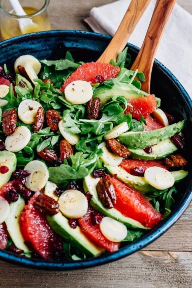 Serving spoons in a bowl of a festive winter grapefruit avocado salad with peppery arugula, hearts of palm, candied pecans, dried cranberries and a maple apple cider vinegar dressing.