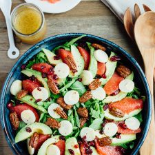 A festive winter grapefruit avocado salad with peppery arugula, hearts of palm, candied pecans, dried cranberries and a maple apple cider vinegar dressing. A plate of grapefruit, serving spoons, and cup of dressing are on the side.