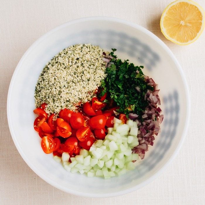 Mixing bowl with the ingredients to make hemp heart tabbouleh: hemp hearts, parsley, red onion, cherry tomatoes and cucumber. A lemon half is beside the bowl