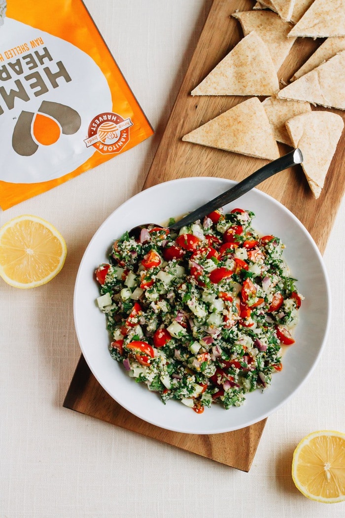 Shallow bowl filled with hemp heart tabbouleh made with hemp, cucumbers, herbs and tomatoes. Beside the bowl are lemon halves, a bag of hemp hearts and pita triangles.