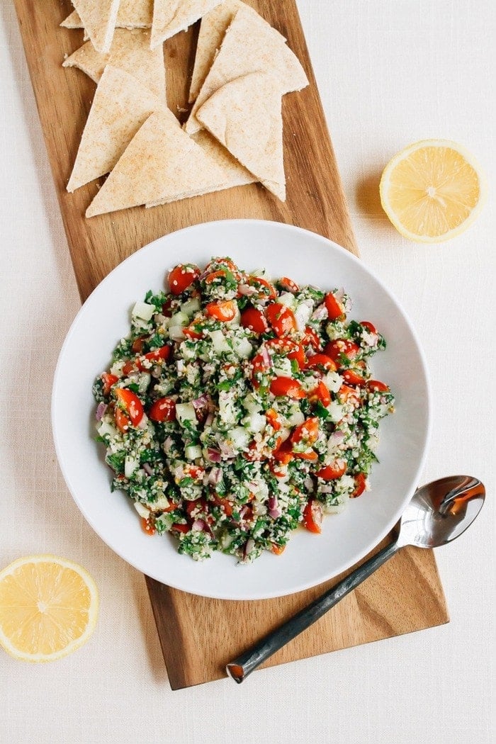 Shallow bowl filled with hemp heart tabbouleh made with hemp, cucumbers, herbs and tomatoes. Beside the bowl are lemon halves, a spoon and pita triangles.