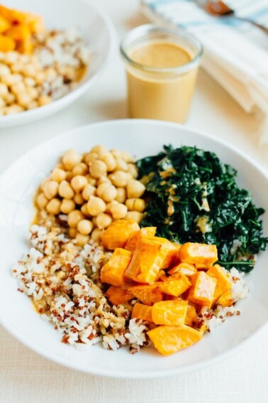 White shallow bowl of brown rice, roasted sweet potato, chickpeas and kale. Drizzled with peanut sauce. The jar of sauce is off to the right-hand side.