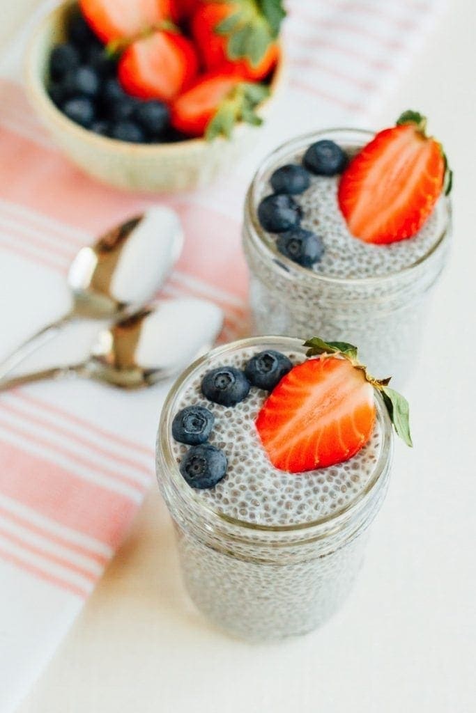 Glass jars with chia pudding, topped with blueberries and strawberries. Napkin, bowl of berries and spoons are to the side.