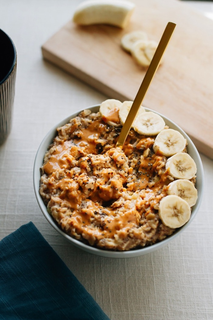 Bowl of oatmeal with chopped bananas arranged on one side of a bowl, peanut butter drizzled on top and a gold spoon sticking out of the bowl. 