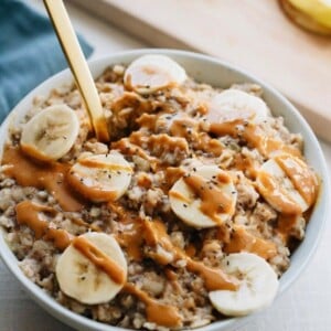 Bowl of oatmeal with chopped bananas and drizzled peanut butter on top. Gold spoon sticking out of the oatmeal. Cutting board with chopped banana out of focus in the background.