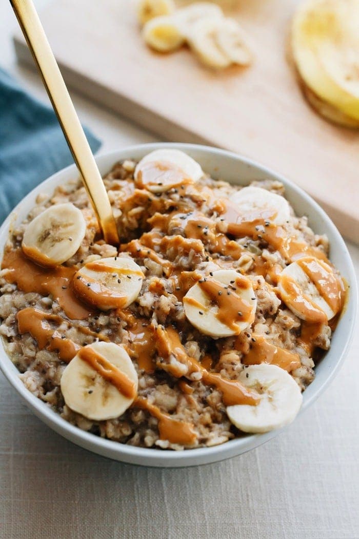 Bowl of oatmeal with chopped bananas and drizzled peanut butter on top. Gold spoon sticking out of the oatmeal. Cutting board with chopped banana out of focus in the background. 