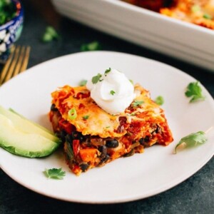 Slice of sweet potato black bean enchilada bake layered with tortillas, sweet potatoes, black beans, cheese and a homemade enchilada sauce with slices of avocado on the side. Remaining enchilada bake in baking dish behind.