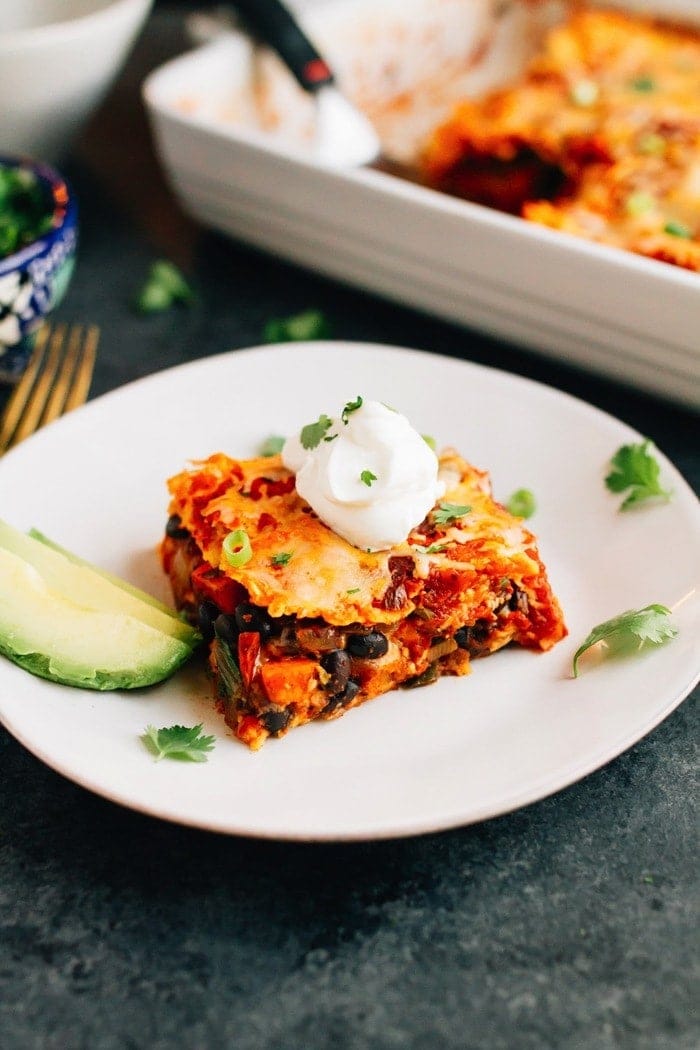 Slice of sweet potato black bean enchilada bake layered with tortillas, sweet potatoes, black beans, cheese and a homemade enchilada sauce with slices of avocado on the side. Remaining enchilada bake in baking dish behind.