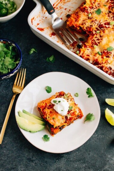 Slice of sweet potato black bean enchilada bake layered with tortillas, sweet potatoes, black beans, cheese and a homemade enchilada sauce with slices of avocado on the side. Remaining enchilada bake in baking dish behind.