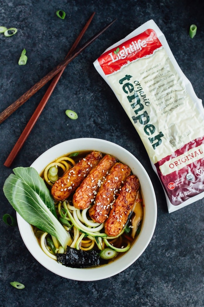 Overhead shot of vegan zucchini noodle ramen bowls served in a white bowl with wood chopsticks on dark grey countertop with Lightlife Tempeh.