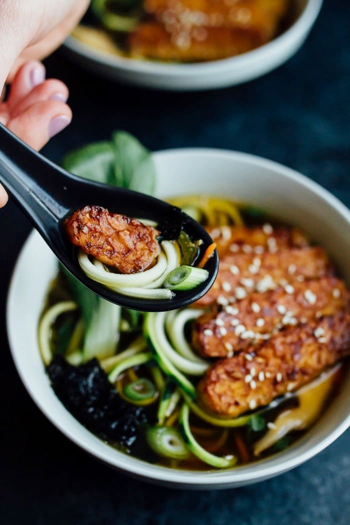 Hand holding black spoon of tempeh and zucchini noodles, over a bowl of vegan zucchini ramen.
