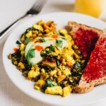 Turmeric tofu scramble on a plate with avocado on top. Two slices of bread with jam on the side. Glass of orange juice and fork also on the side.