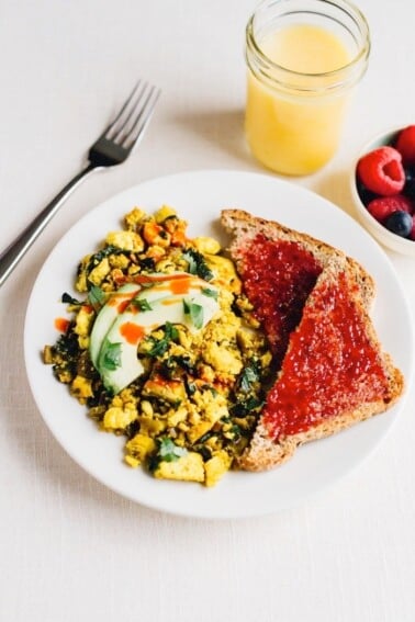 Turmeric tofu scramble on a plate with avocado on top. Two slices of bread with jam on the side. A bowl of raspberries and blueberries is next to the plate. Glass of orange juice and fork also on the side.