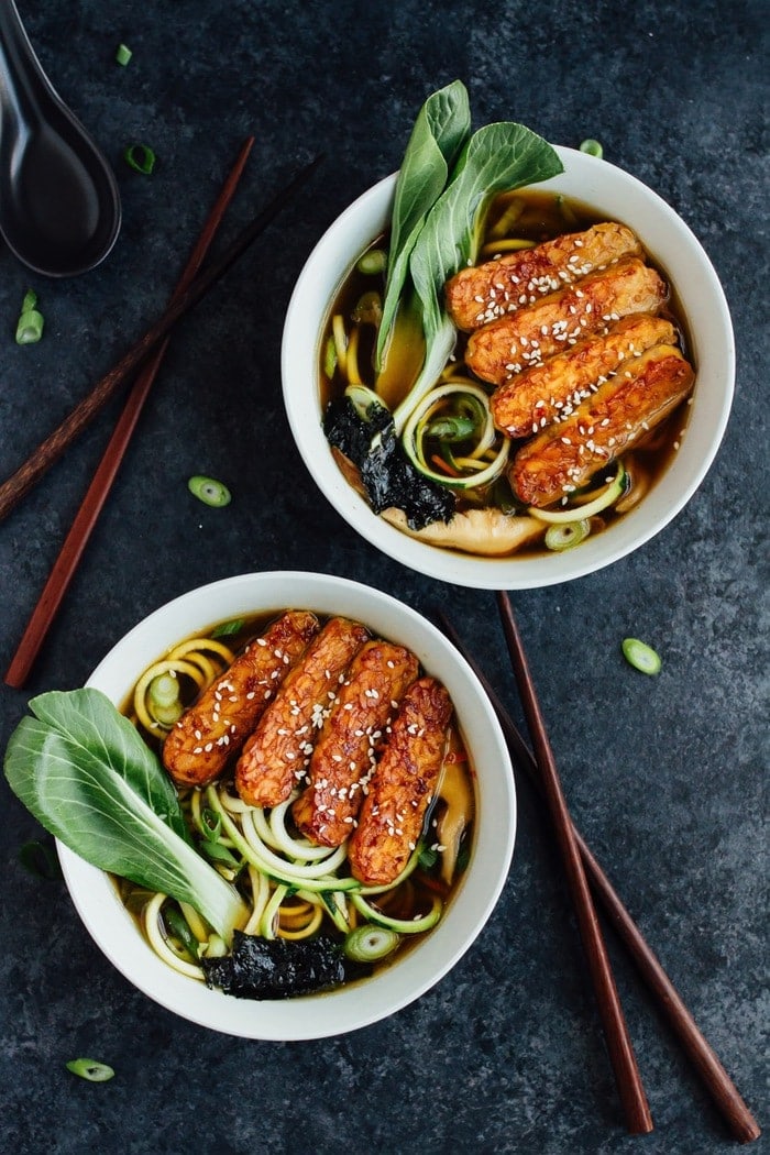 Overhead shot of vegan zucchini noodle ramen bowls served in white bowls with wood chopsticks on dark grey countertop.