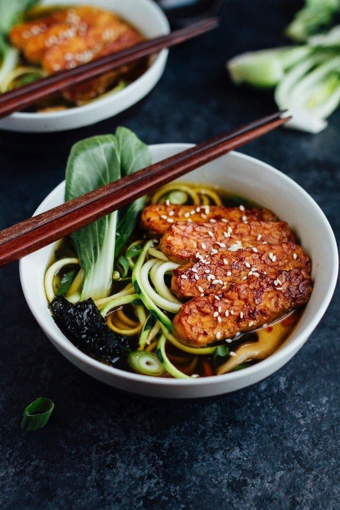 Vegan zucchini noodle ramen bowls served in white bowls with wood chopsticks on dark grey countertop.