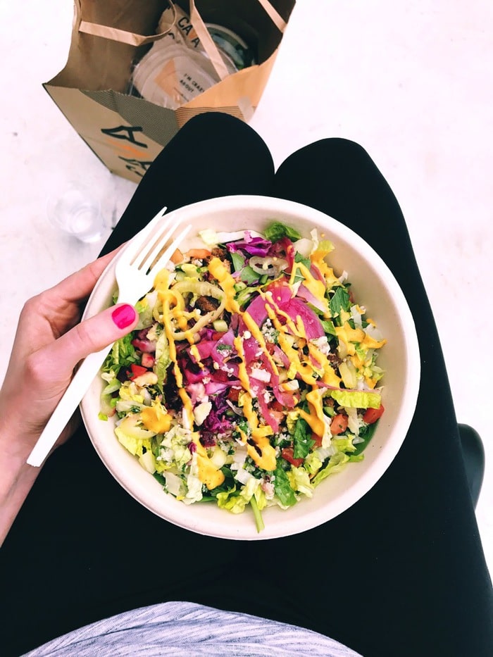 Cava Grill.jpg A woman holding a salad in her lap with a fork.