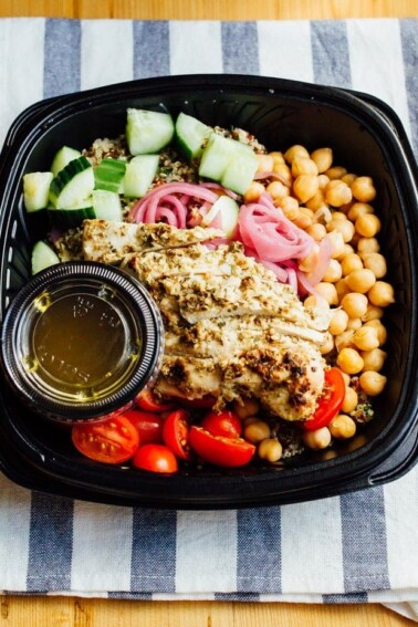 Stellas greek chicken quinoa bowl in a to-go container with cucumber, tomatoes, chicken, purple onion and chickpeas, sitting on a blue and white striped towel.