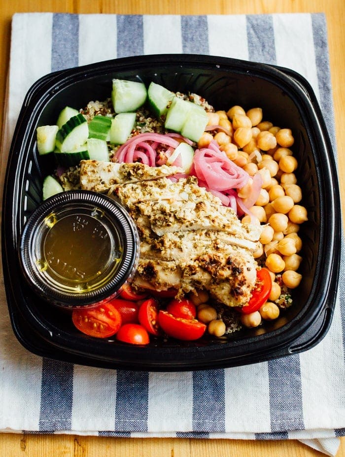 Stellas greek chicken quinoa bowl in a to-go container with cucumber, tomatoes, chicken, purple onion and chickpeas, sitting on a blue and white striped towel.