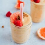 Two grapefruit pineapple smoothies with pink straws, topped with granola, a raspberry and a slice of grapefruit. They are surrounded by raspberries and slices of grapefruit on the counter.