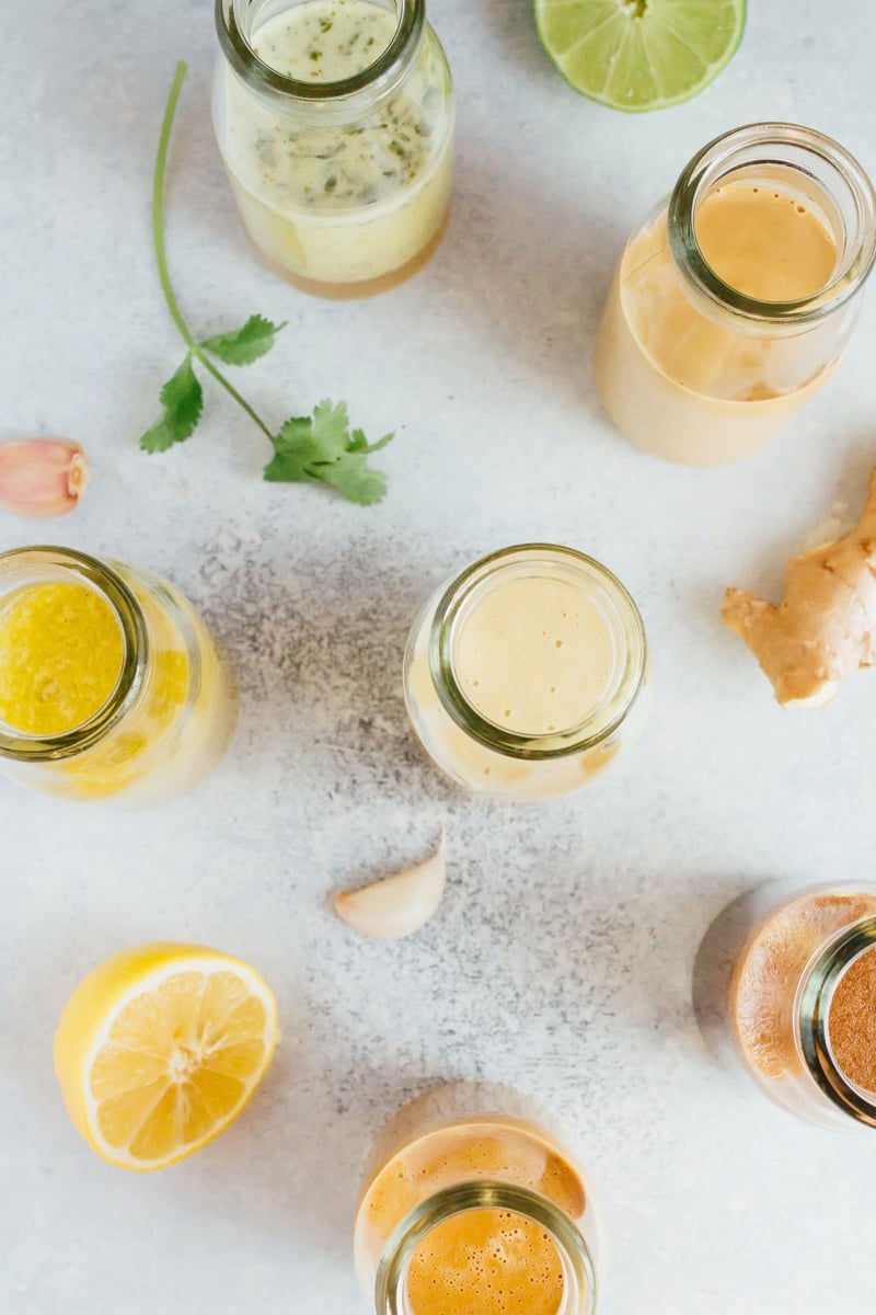 Healthy homemade salad dressings in a glass jars spread across a counter top with fresh garlic, lime, ginger and cilantro in the background. 