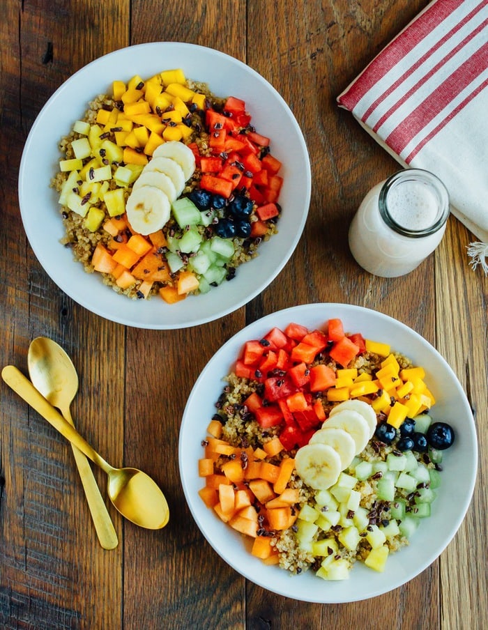 Overhead shot of Cinnamon Quinoa Breakfast Bowls with chopped fresh fruit, served in white bowls on wood table.