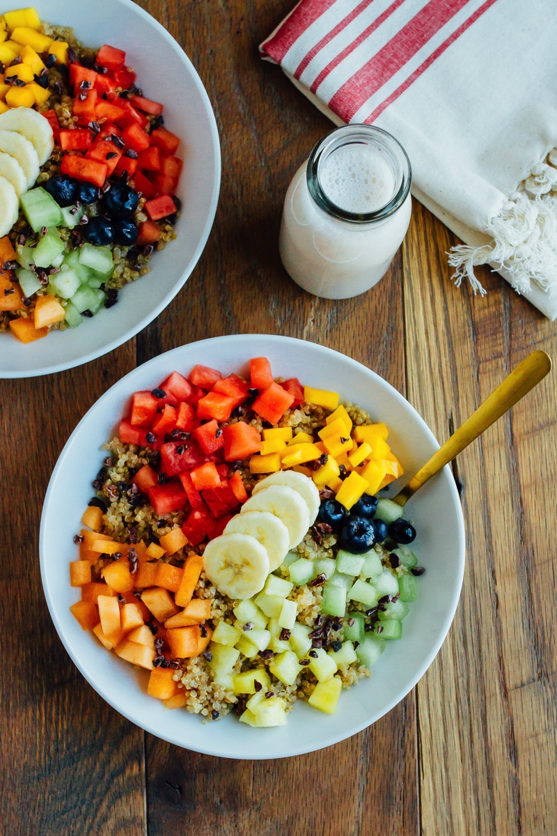 Overhead shot of Cinnamon Quinoa Breakfast Bowls with chopped fresh fruit, served in white bowls on wood table.