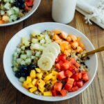 Overhead shot of Cinnamon Quinoa Breakfast Bowls with chopped fresh fruit, served in white bowls on wood table and glass of milk.