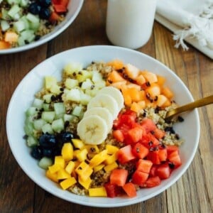 Overhead shot of Cinnamon Quinoa Breakfast Bowls with chopped fresh fruit, served in white bowls on wood table and glass of milk.