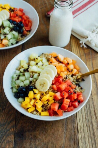 Overhead shot of Cinnamon Quinoa Breakfast Bowls with chopped fresh fruit, served in white bowls on wood table and glass of milk.