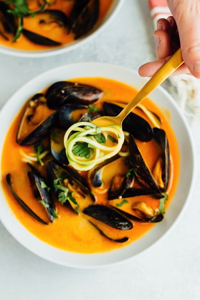 Hand holding a spoon of the coconut curry broth with zucchini noodles. Bowl of mussels in the background.