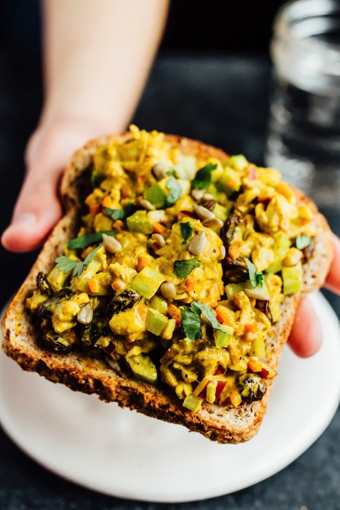 Curried tempeh salad on a slice of bread a hand is holding. Salad includes sunflower seeds, curry dressing, raisins and veggies.