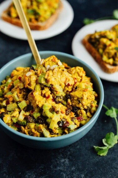 Curried tempeh salad in a serving bowl next to two plates with the salad on bread.