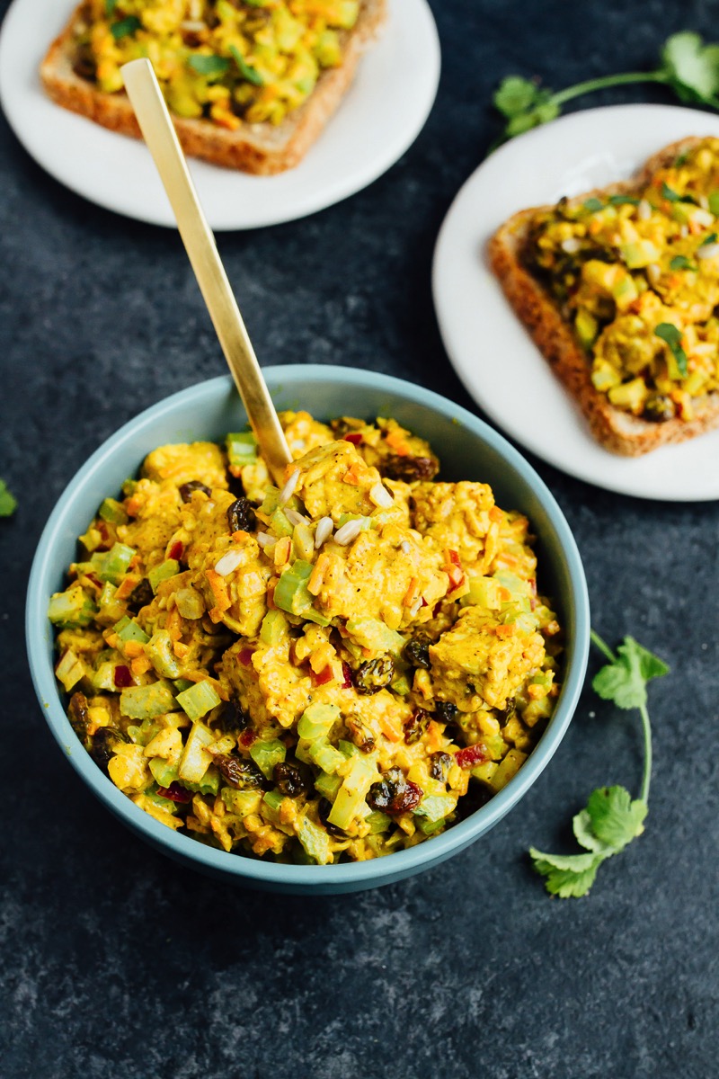 Curried tempeh salad with celery and raisins in a bowl and on two sliced of bread.