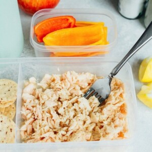 A to-go container full of tuna with a fork in it, sliced peppers in a container in the background.