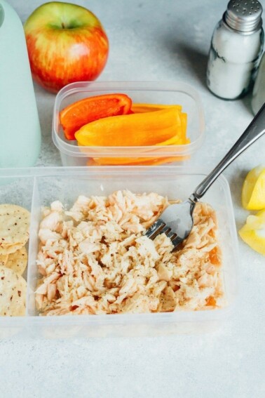 A to-go container full of tuna with a fork in it, sliced peppers in a container in the background.