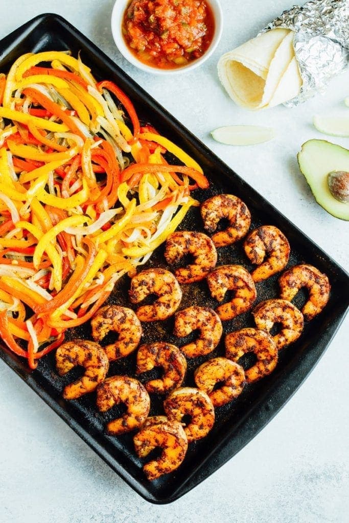 Sheet pan with baked shrimp coated in Mexican spices, peppers and onions. The table is topped with a bowl of salsa, tortillas, lime and avocado.