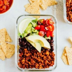 A glass dish with tempeh salad, including tempeh, black beans, lettuce tomatoes, and lime. Salsa, tempeh, and chips on the table on the side.