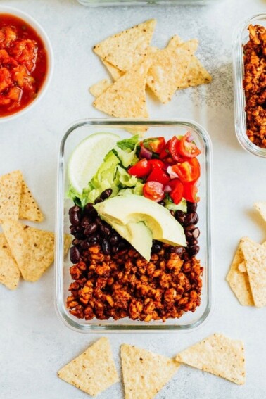 A glass dish with tempeh salad, including tempeh, black beans, lettuce tomatoes, and lime. Salsa, tempeh, and chips on the table on the side.