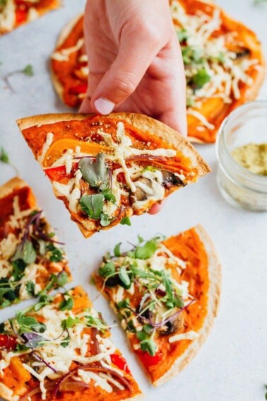 Hand holding vegan hummus tortilla pizza with peppers, greens, mushrooms and vegan cheese.