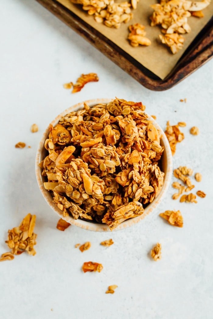 Bowl of granola surrounded by granola on the table, baking sheet in the background.