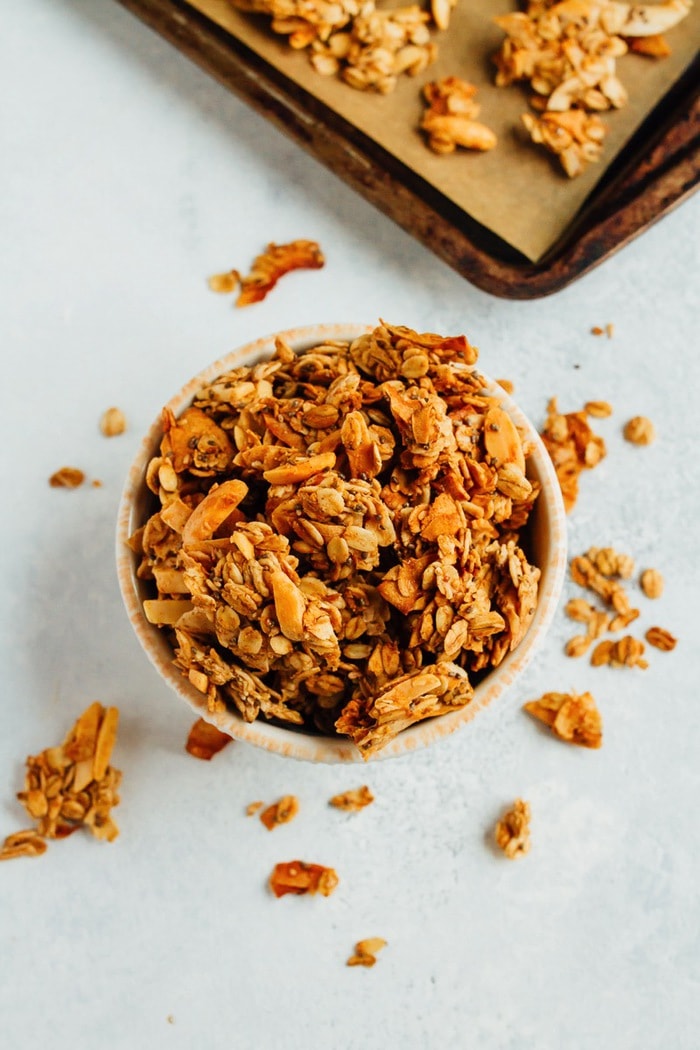 Bowl of granola surrounded by granola on the table, baking sheet in the background.