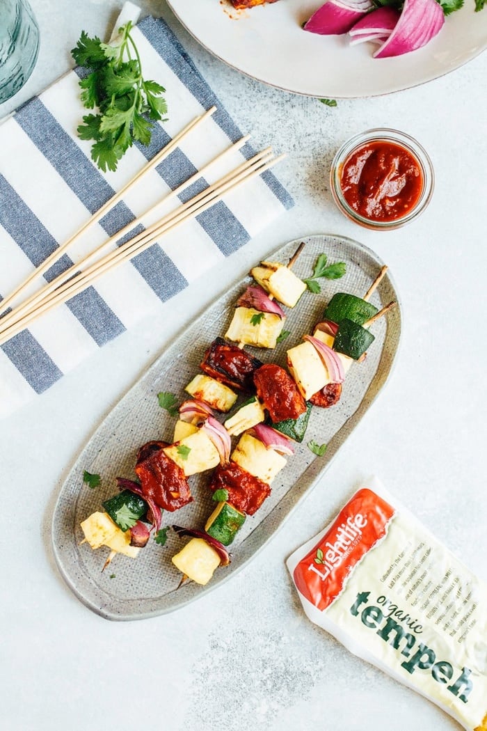 Three bbq tempeh skewers with onion, pineapple and zucchini on a plate.