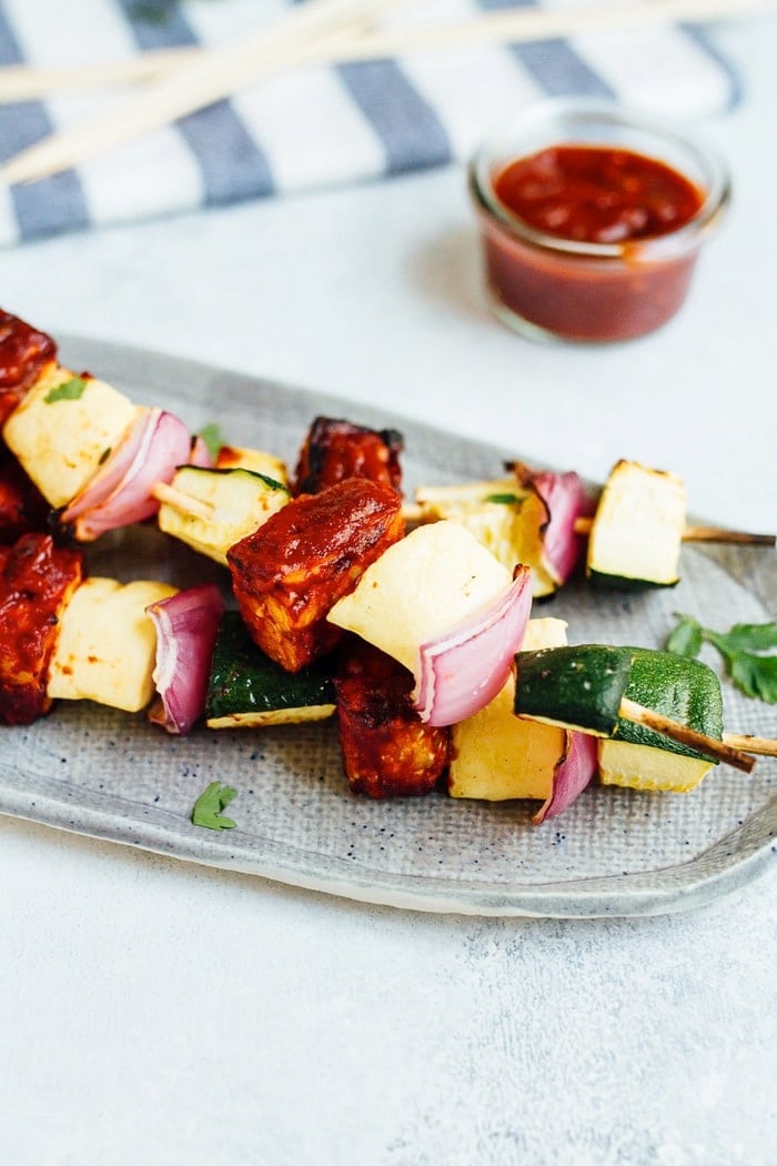 Three bbq tempeh skewers with onion, pineapple and zucchini on a plate.