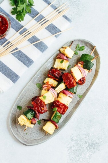 Grilled BBQ tempeh and veggie skewers on an oval platter, next to skewers on a towel.