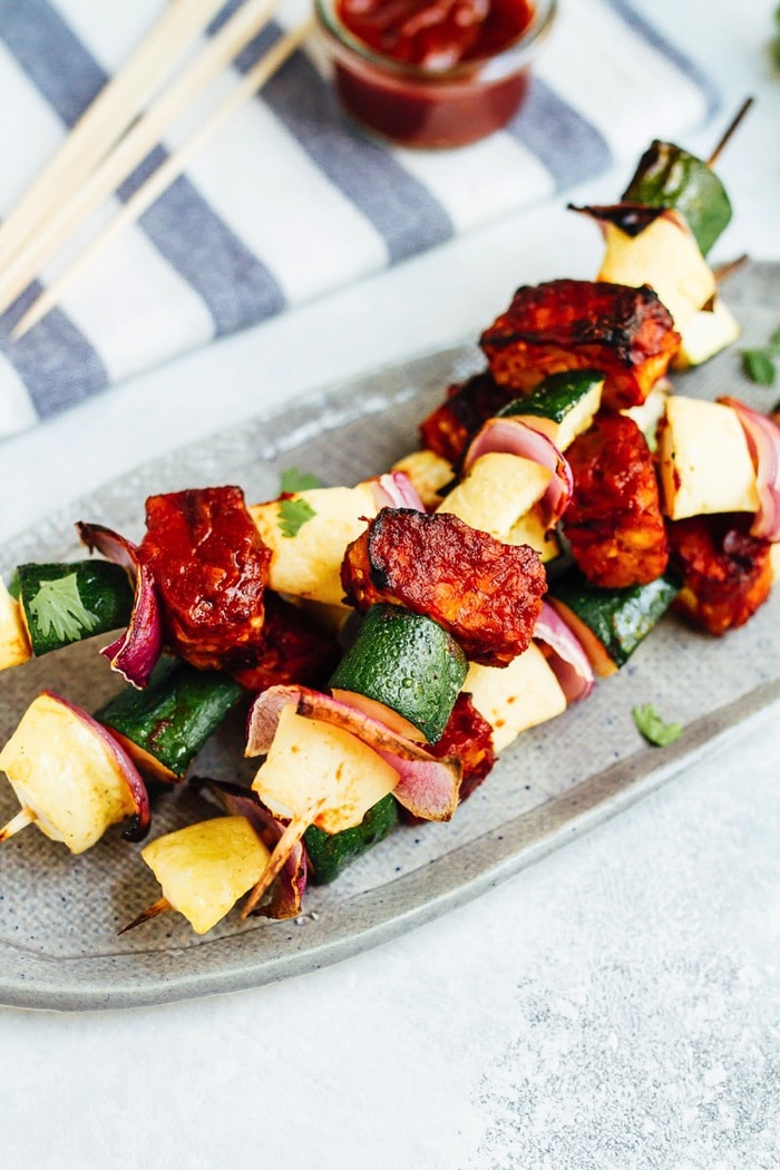 Three bbq tempeh skewers with onion, pineapple and zucchini on a plate.