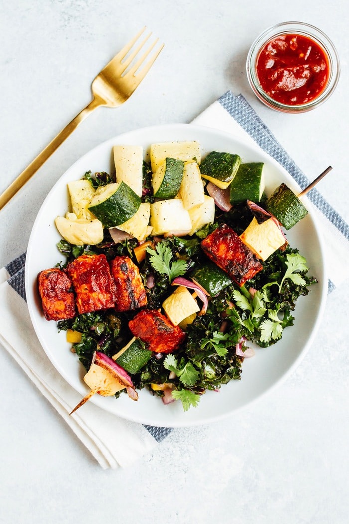 Bbq tempeh skewers with onion, pineapple, and zucchini on a salad with a dish of bbq sauce next to the plate.