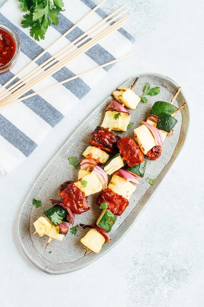 Three bbq tempeh skewers with onion, pineapple and zucchini on a plate.